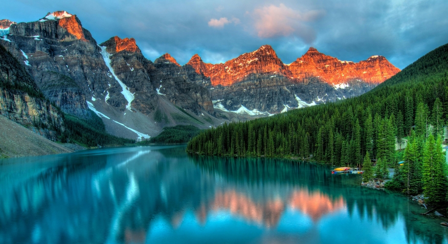 Mountain range with peaks glowing in the sunset, mirrored in a clear, calm lake bordered by dense forest.