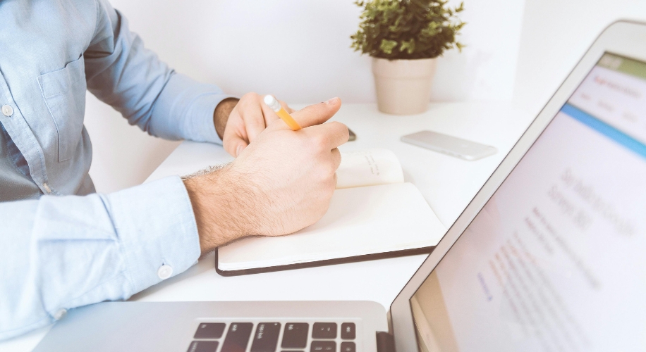 Person writing in a notebook while using a laptop, focusing on the notebook and pencil.