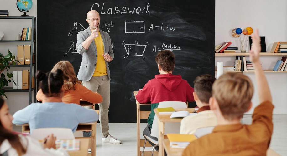 Teacher in front of a blackboard with geometry homework, students raising hands to answer. Text on blackboard: Classwork. A = L × w, b × (h / 2), A = (B + b) × h.