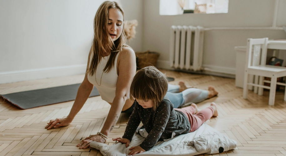 Woman practicing yoga with a child on a white blanket in a room with a wooden floor.