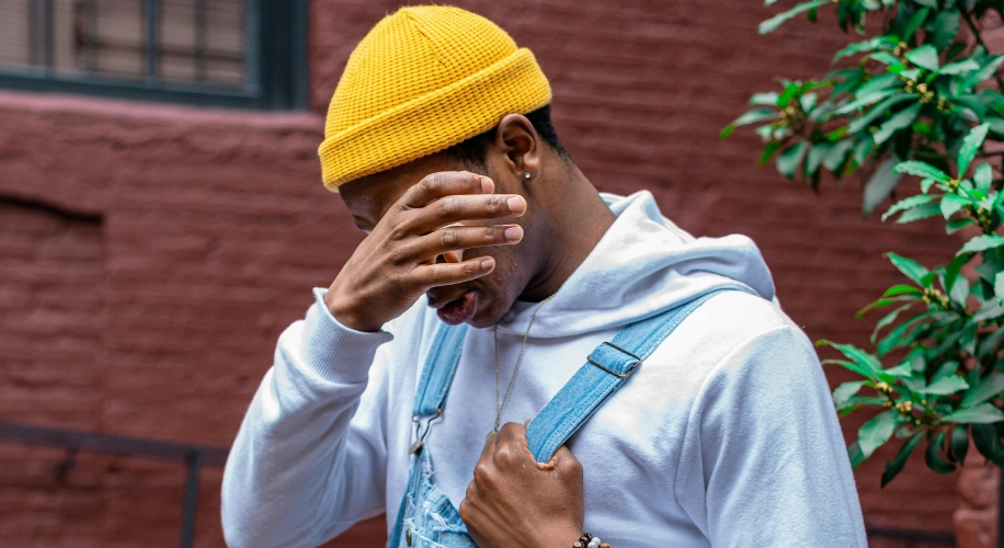 Person wearing a yellow knit beanie, white hoodie, and blue overalls, covering face with hand.