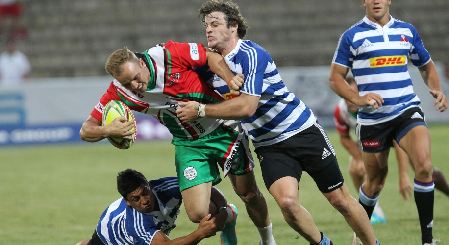 Rugby player in red and green jersey breaking a tackle from two players in blue and white striped jerseys.