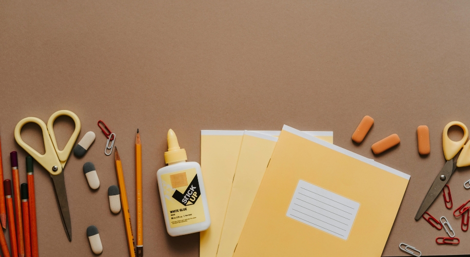 Scissors, pencils, erasers, a bottle of Stick Up white glue, folders, and paper clips on a brown background.