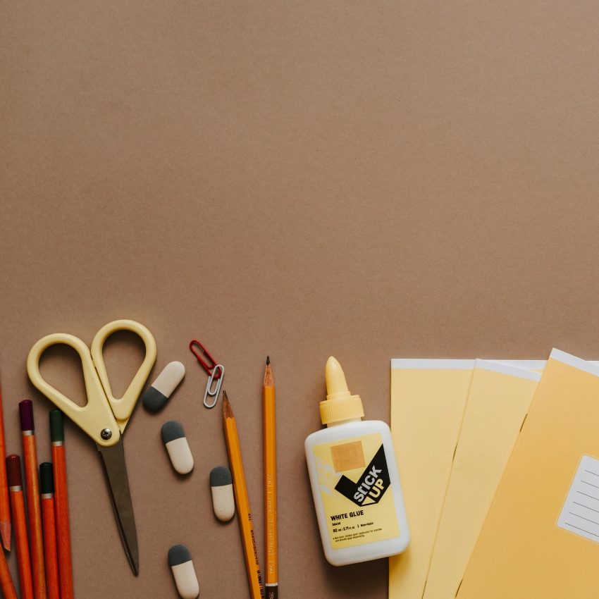 Glue, scissors, pencils, erasers, paperclips, crayons, and yellow envelopes scattered on a brown surface. Glue says "STICK UP, WHITE GLUE, 40 ml., 1.35 FL oz, Non-toxic Formula."