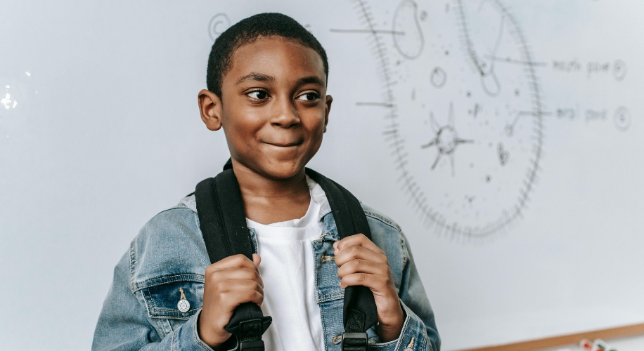 Young boy smiling, wearing a denim jacket and holding a black backpack strap.