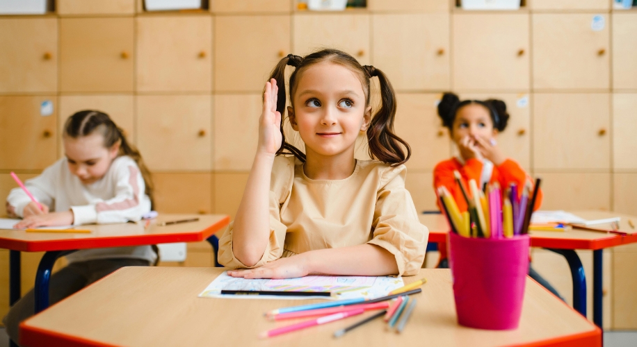 Child raising hand in classroom, sitting at a desk with colorful pencils and markers on the table.