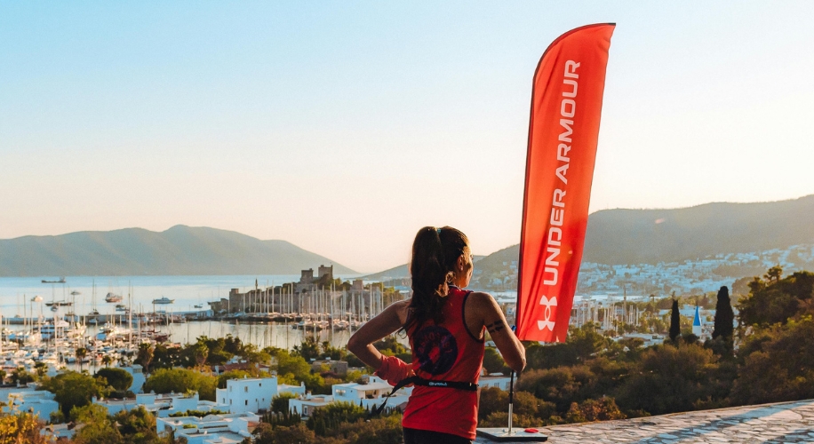 A person in a red Under Armour tank top standing next to an Under Armour flag overlooking a marina.