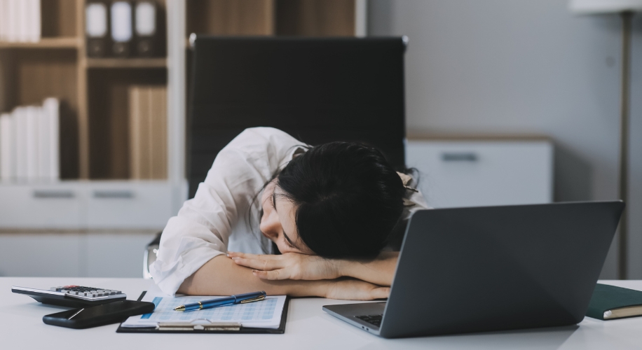 Person resting their head on a desk with a laptop, calculator, pens, and documents in an office setting.