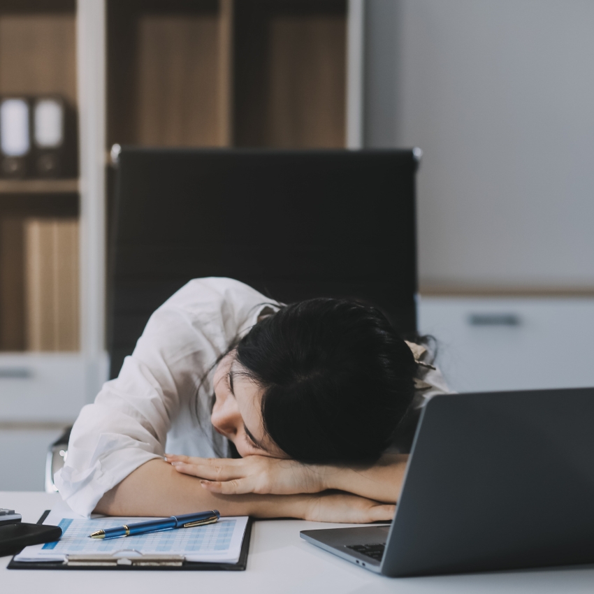 Woman asleep at a desk with an open laptop, clipboard with papers, calculator, and pens nearby.