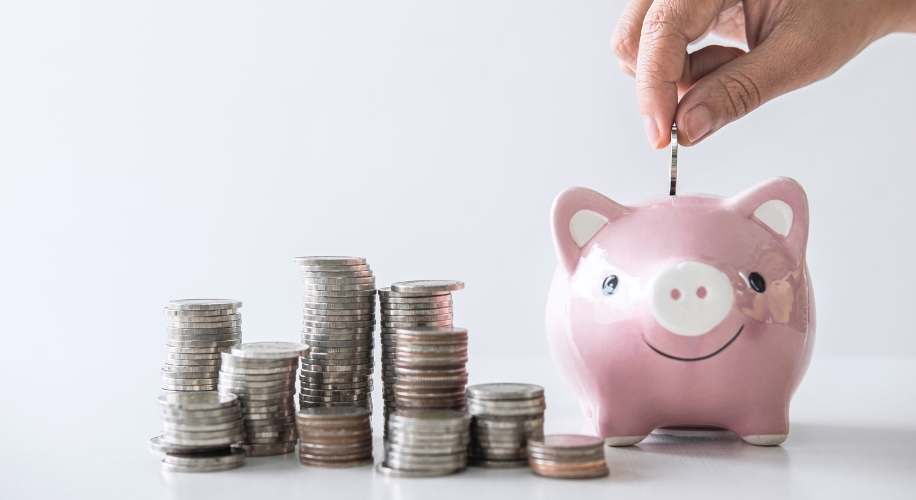 Hand placing a coin into a smiling pink piggy bank next to stacks of coins.