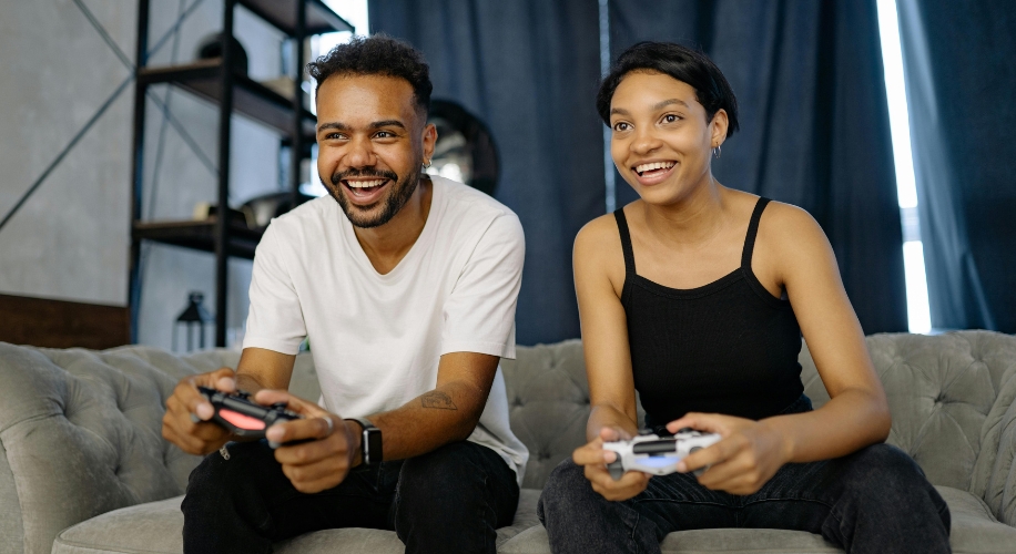 Two people smiling while holding game controllers, sitting on a gray couch.