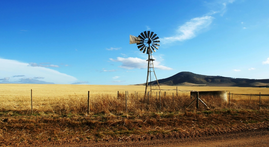 Windmill standing in a vast dry field with a mountain in the background under a clear blue sky.