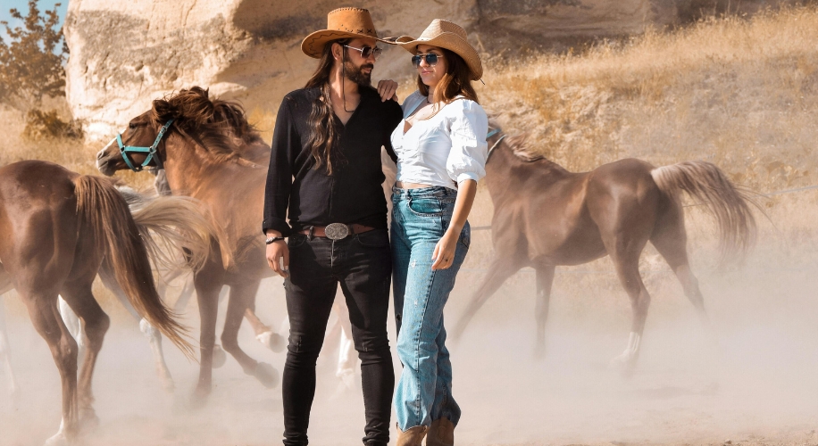 Two people wearing cowboy hats standing together in front of a group of horses.