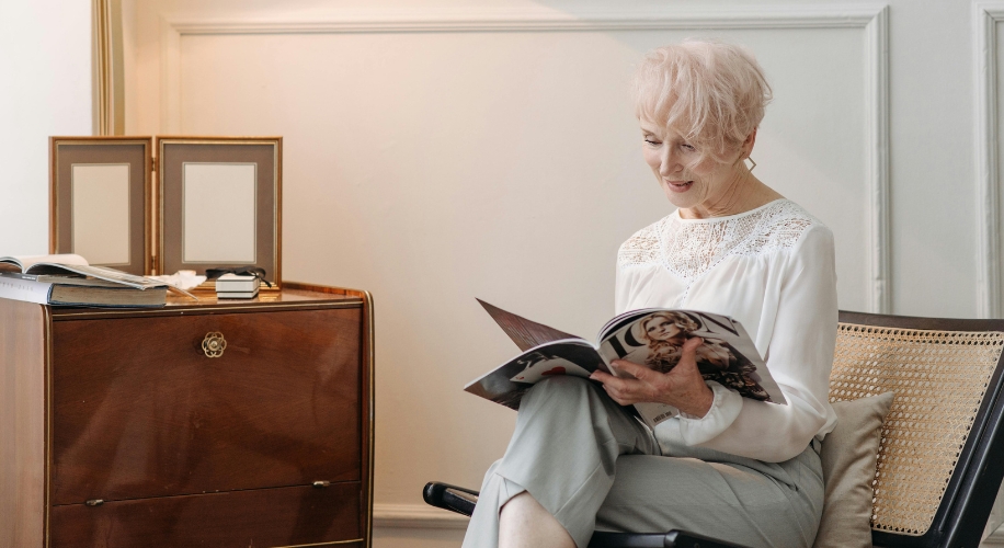 Senior woman reading a fashion magazine in a light-colored room, sitting on a chair with a cushion.