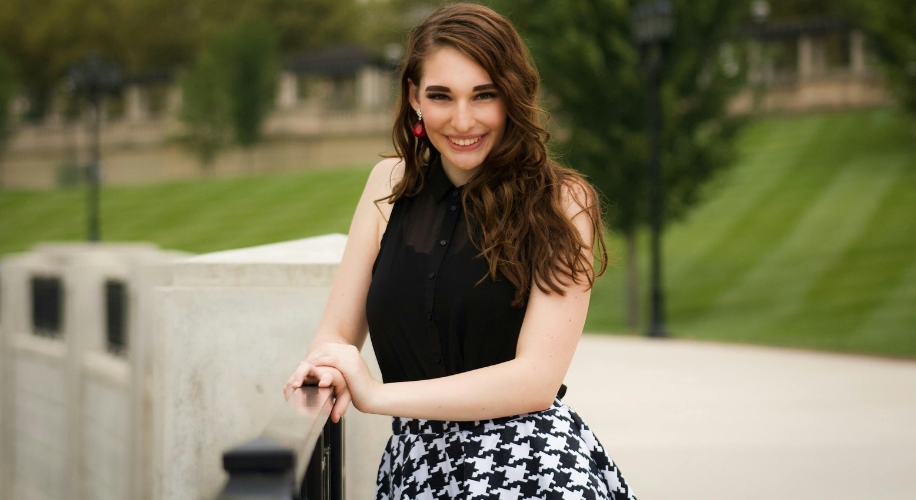 Woman wearing a black sleeveless top and a black-and-white houndstooth skirt, smiling while leaning on a railing.