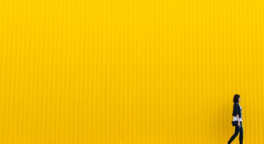 A person in black jacket and blue jeans walking in front of a large, yellow corrugated metal wall.