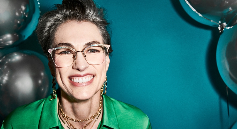 Smiling individual wearing geometric eyeglasses with a gold bridge, dressed in a green shirt with layered gold necklaces and dangling earrings.