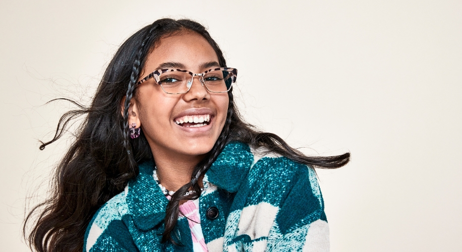 Young girl wearing leopard-print eyeglasses and a teal and white plaid jacket, smiling brightly.