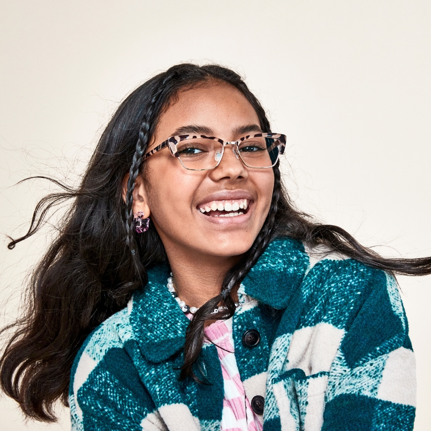 Young girl wearing tortoiseshell eyeglasses and a teal plaid jacket, smiling broadly.