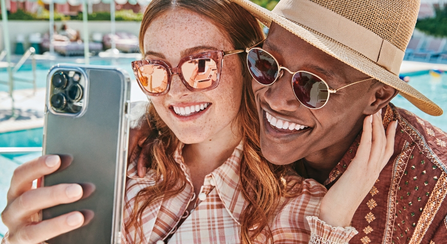 Close-up of two people wearing sunglasses, with one holding a smartphone to take a selfie by the pool.