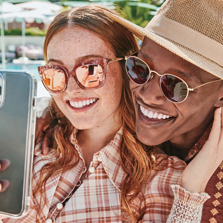 Two people wearing stylish sunglasses and smiling while taking a selfie with a smartphone.