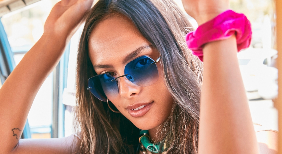 Woman wearing aviator sunglasses with blue lenses and a pink scrunchie on her wrist.