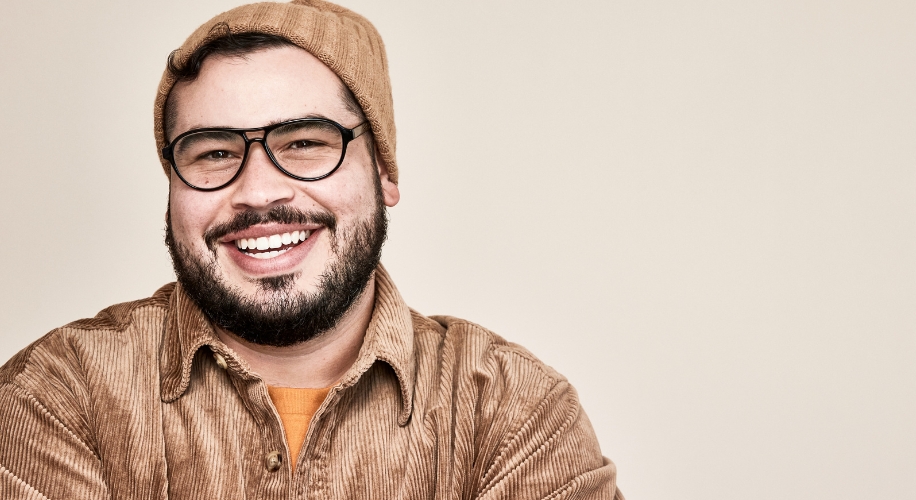 Man wearing glasses, a brown beanie, and a brown corduroy jacket, smiling against a plain background.