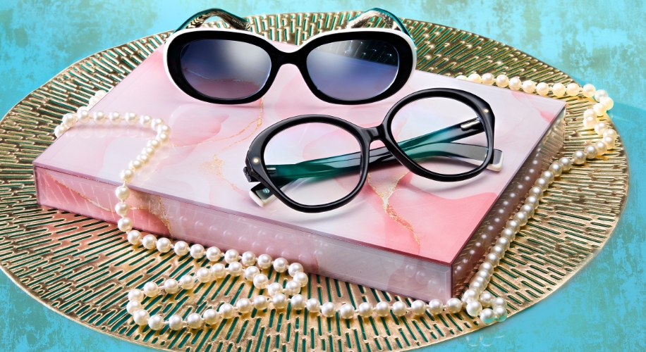 Two pairs of glasses, one with black and white frames, one with round black frames, on a pink book with pearl necklaces.