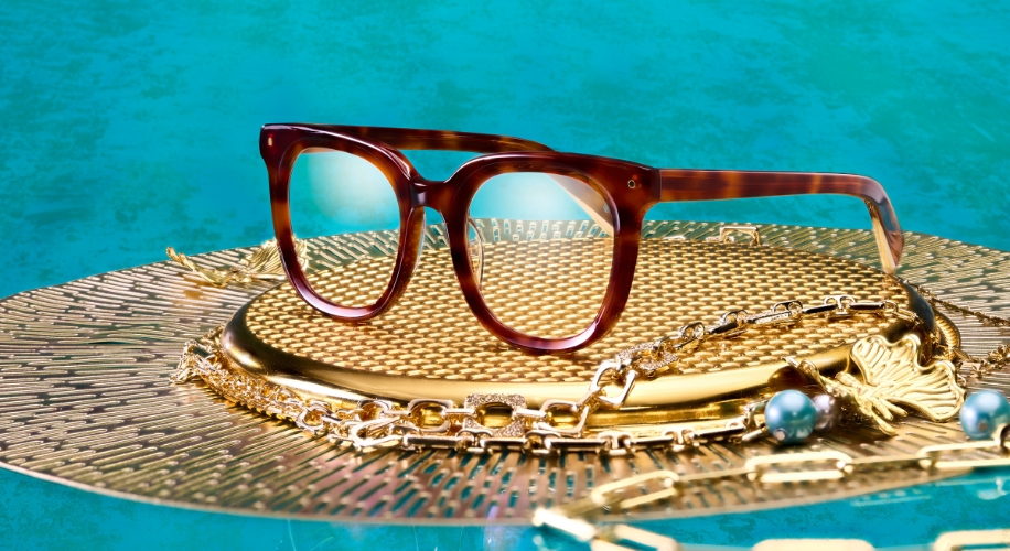 Tortoiseshell eyeglasses with thick frames, displayed on a gold textured surface surrounded by jewelry.