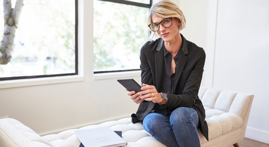 Person holding a tablet, seated on a beige cushioned bench, with a notebook and glasses on the bench.