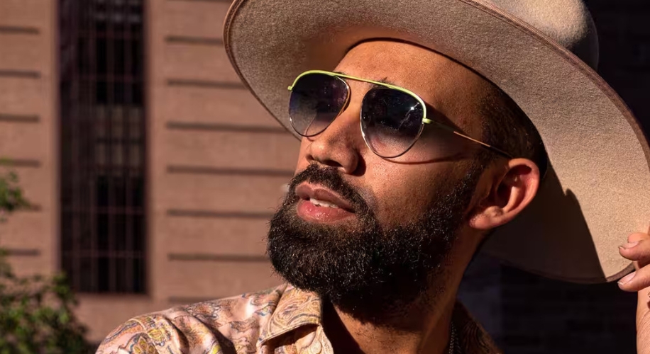 Wide-brimmed beige hat and gold-framed sunglasses with black lenses on a person with a beard.