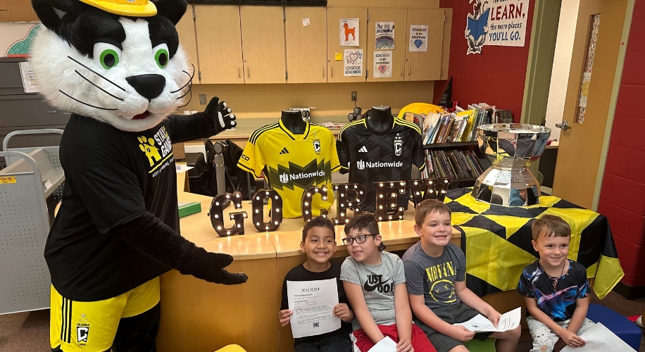 Mascot pointing at Columbus Crew merchandise including jerseys, a GO CREW sign, and a silver trophy.