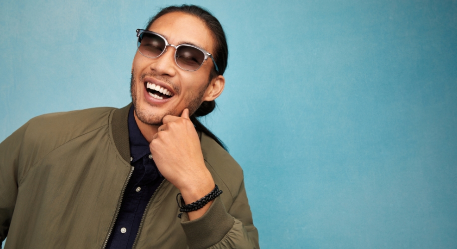 Man wearing clear-framed sunglasses, olive bomber jacket, dark shirt, and bracelet, smiling against a blue background.