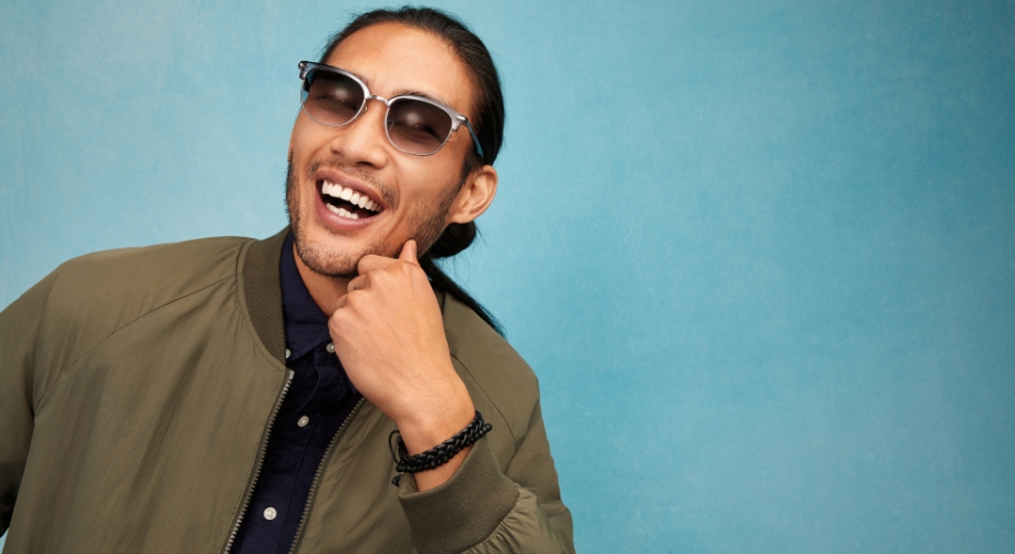 Man wearing stylish sunglasses and a green jacket, smiling against a blue background.