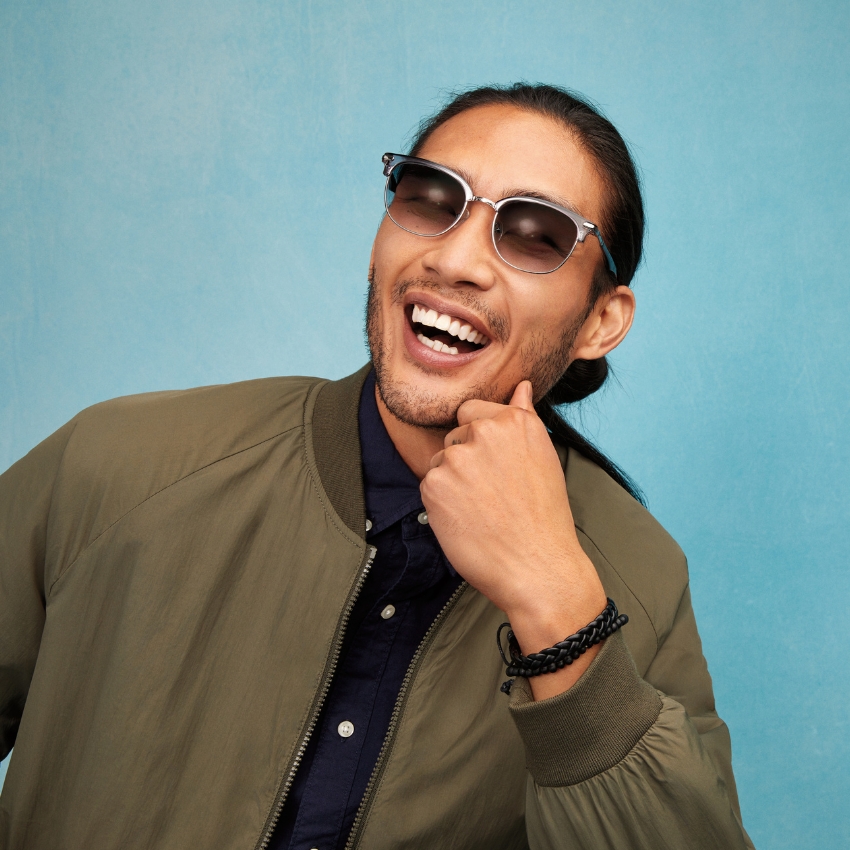 Man wearing sunglasses and a khaki jacket, smiling with hand on chin.