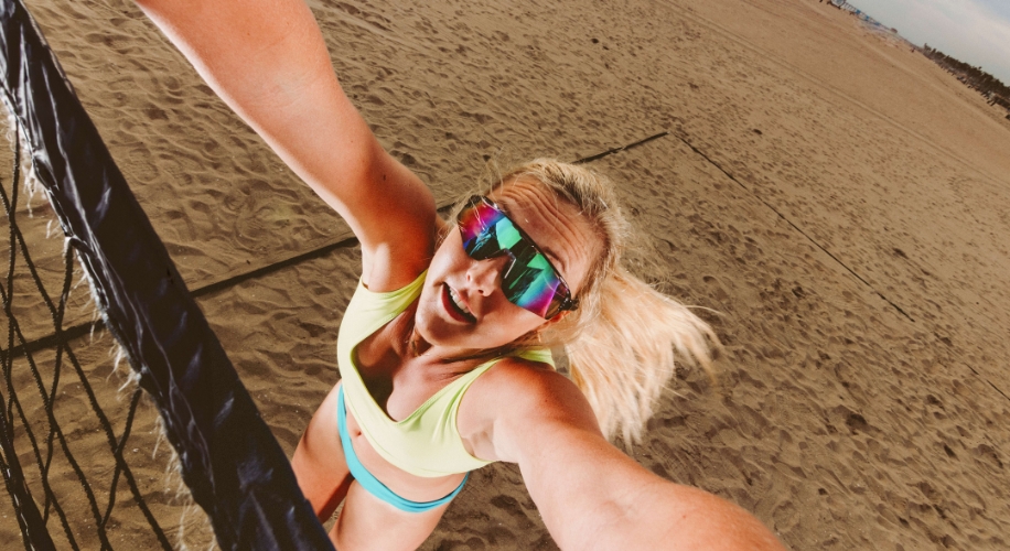 Woman in vibrant sports attire wearing mirrored sunglasses, reaching up to hit a volleyball.