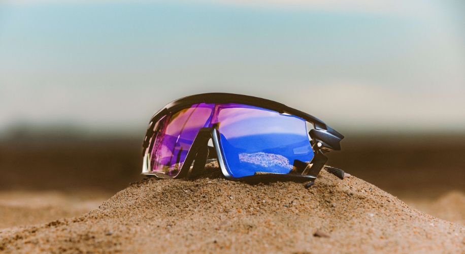 Polarized sport sunglasses with purple and blue lenses resting on sand.