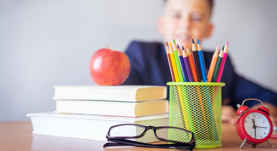 Stack of books with an apple on top, next to colored pencils in a holder, eyeglasses, and a red alarm clock.