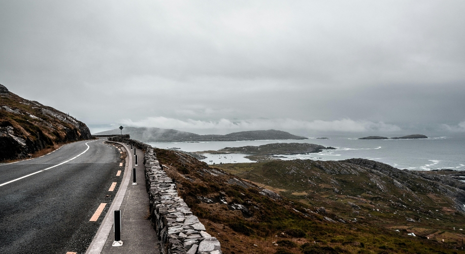 Winding coastal road next to rocky hills and overlooking a foggy ocean with distant islands.