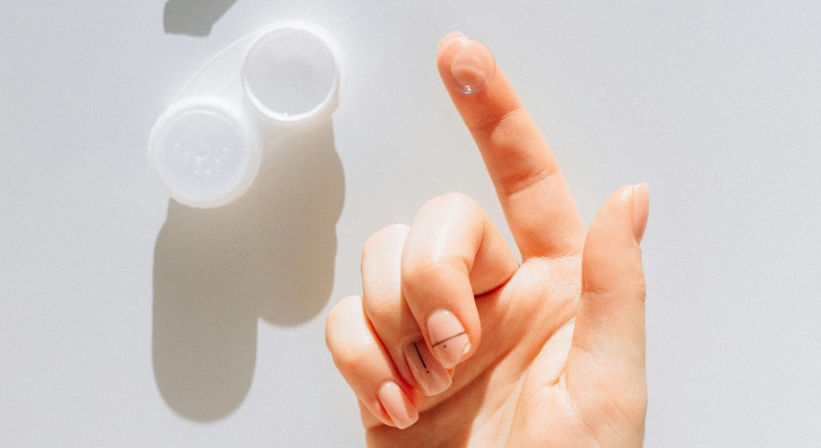 Hand holding a transparent contact lens on a fingertip with a white contact lens case in the background.