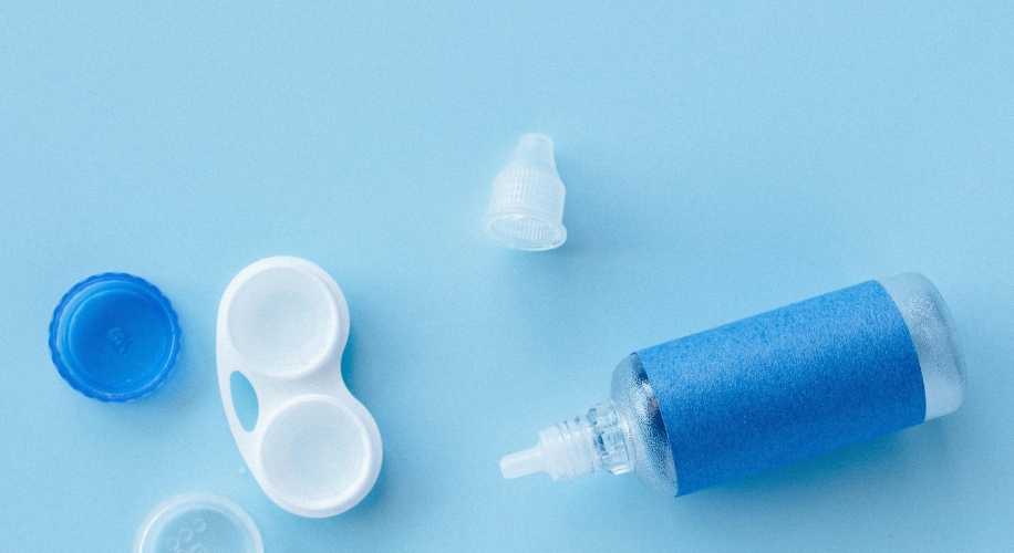 Contact lens solution bottle, open lens case, and blue lid on a light blue surface.