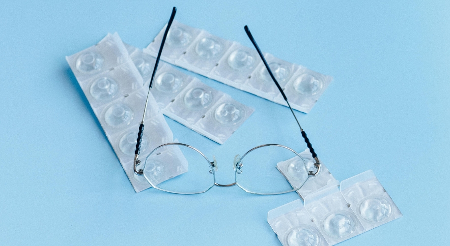 Eyeglasses placed on top of contact lens blister packs on a light blue background.