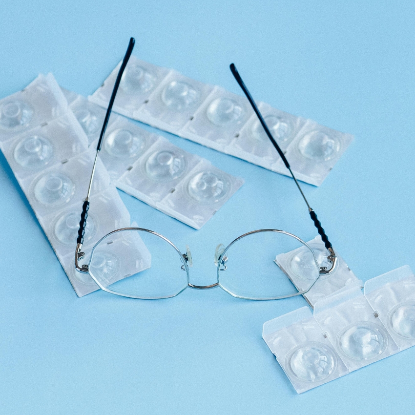 Eyeglasses placed on top of contact lens blister packs on a light blue background.