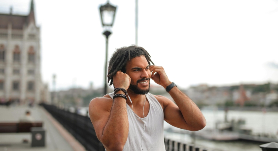 Man wearing wireless earbuds and a light grey sleeveless shirt, smiling and adjusting his earbuds outdoors.