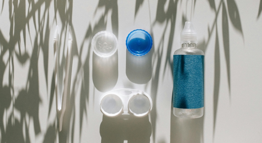 Contact lens case, lens solution bottle with blue label, tweezers, and lens holder on white surface.