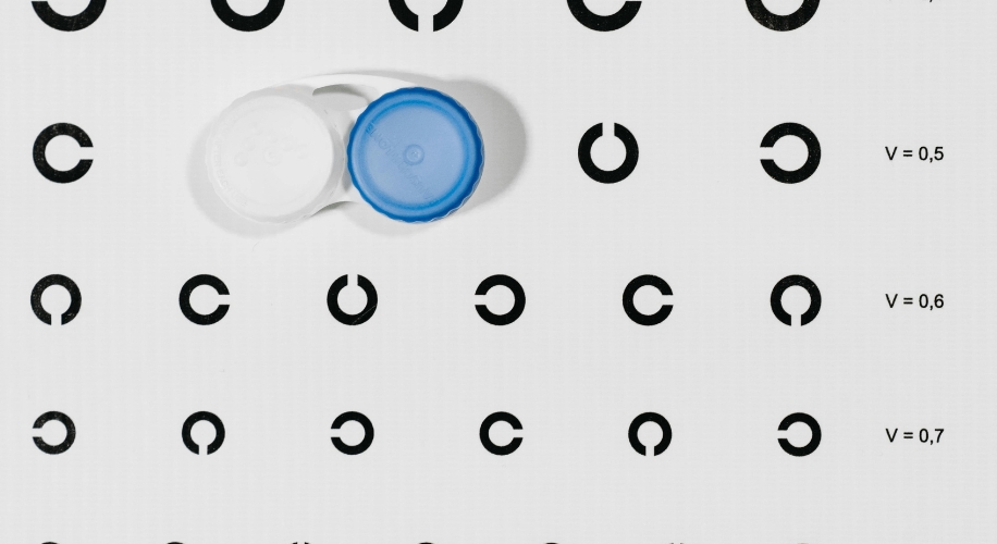 Contact lens case with blue and white caps on an eye chart featuring C-shaped symbols and vision levels V=0.5, V=0.6, V=0.7 on the right.