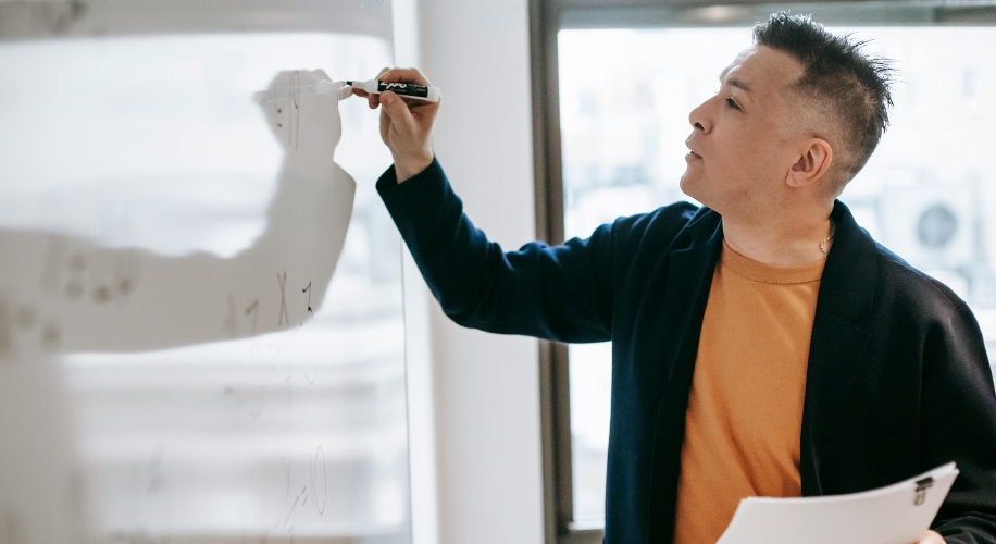 Person writing on a whiteboard with a dry erase marker while holding papers in the other hand.