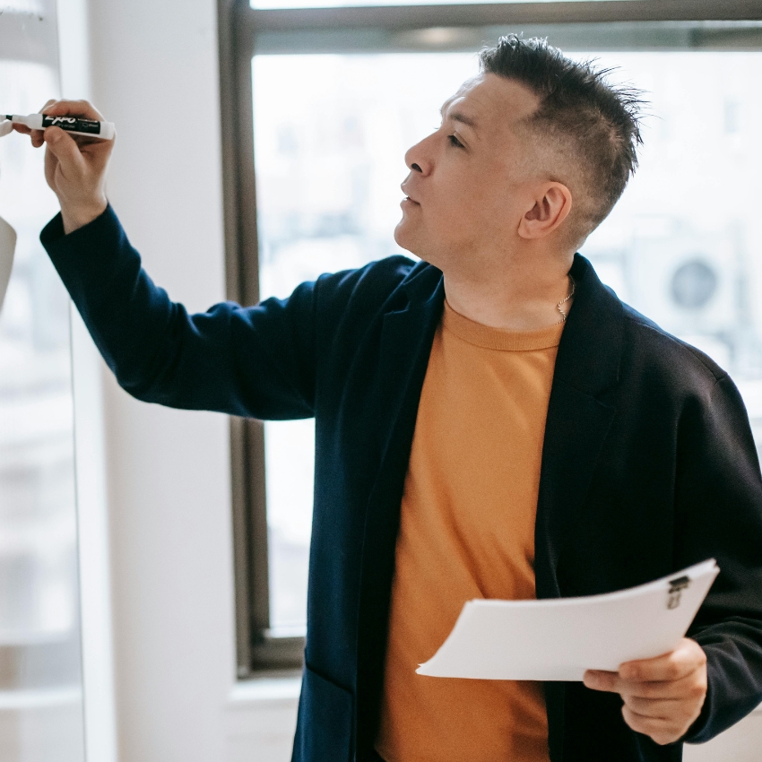 Person writing on a whiteboard with a marker, holding papers in the other hand, dressed in a blazer and shirt.