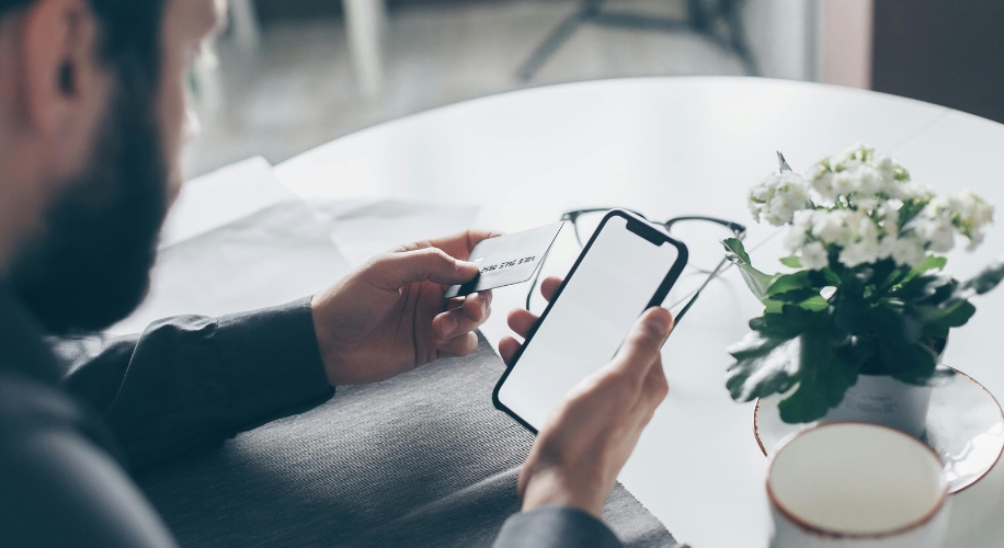 Man holding a smartphone and credit card at a table with white flowers and a coffee mug.