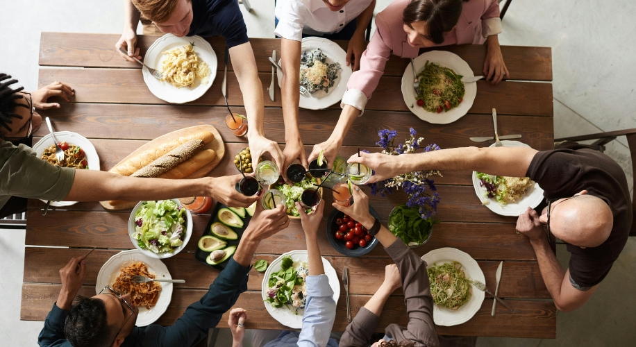 People enjoying different pasta dishes and salads at a wooden table, toasting with drinks.